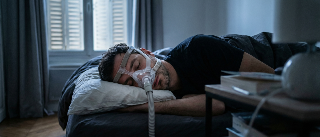 Homme endormi paisiblement dans son lit portant un masque de PPC (Pression Positive Continue) pour traiter son apnée du sommeil.